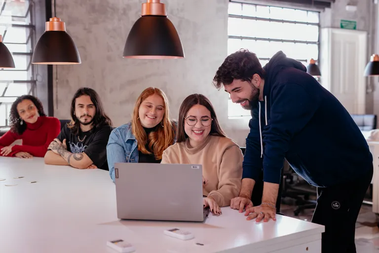 Cinco pessoas do time da Prod reunidas ao redor de um notebook, sorrindo durante uma reunião no escritório.