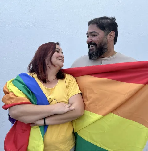 Duas pessoas do time da Prod sorrindo e abraçadas, segurando a bandeira do orgulho LGBTQIA+, em ação do comitê prodiversidade.